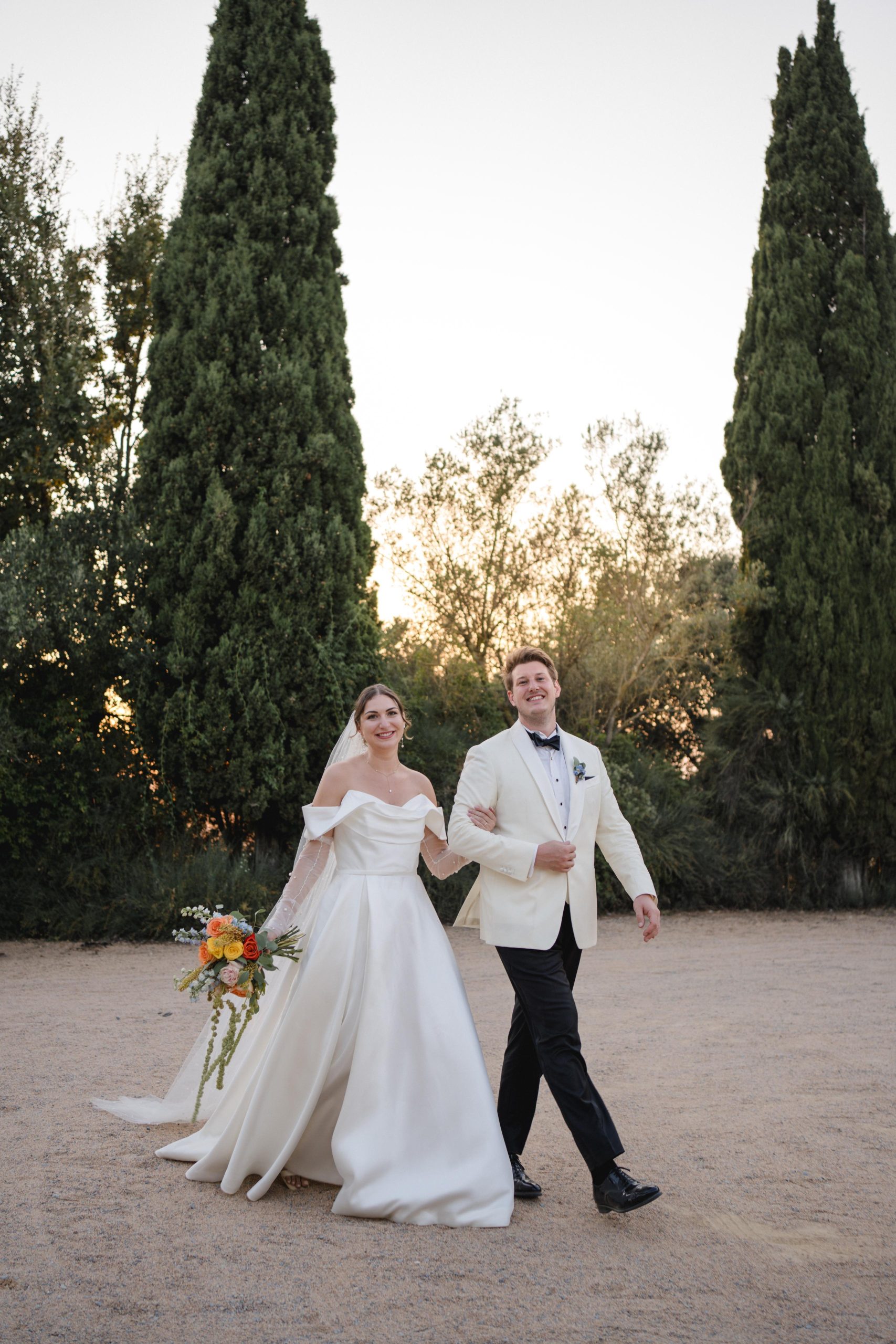 Novia y novio caminando juntos al atardecer en su boda en Barcelona, ella con maquillaje natural y vestido blanco elegante.