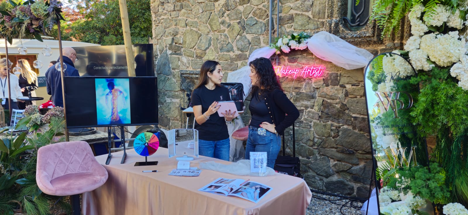 Stand de maquillaje en el Wedding Market en Barcelona, con decoración floral, una mesa con productos de belleza, un espejo iluminado y dos personas conversando en el espacio de Makeup Artist.
