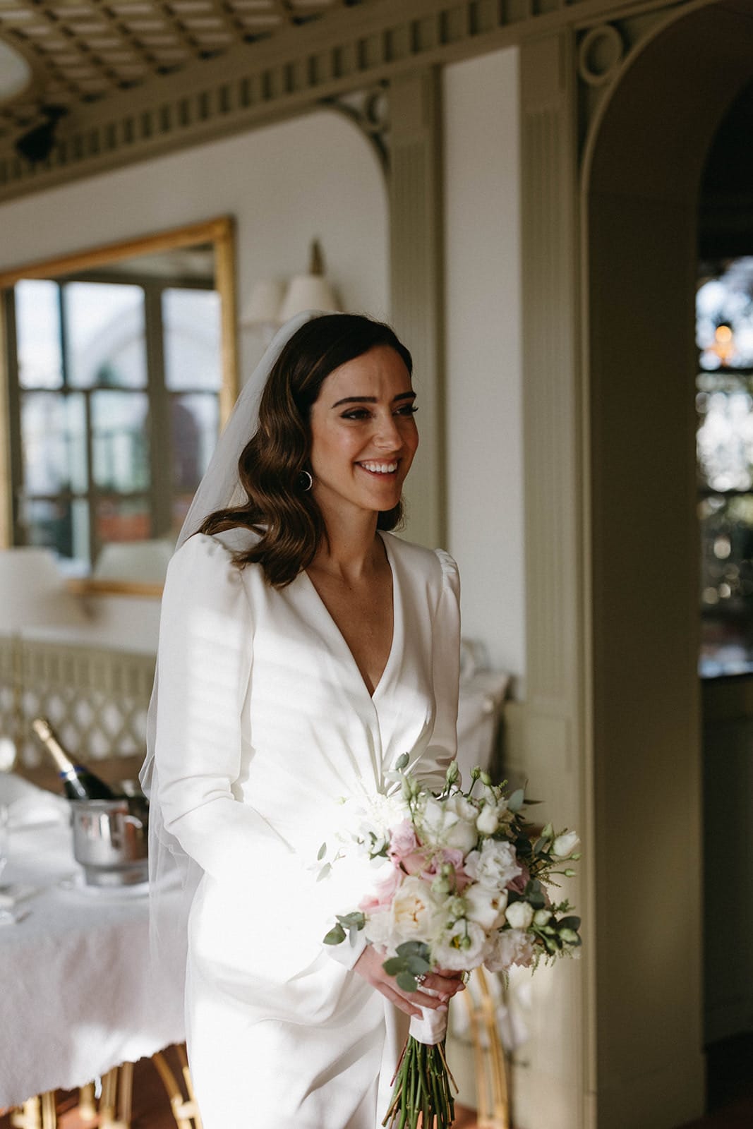 Novia sonriente con maquillaje natural y elegante sosteniendo ramo de flores en interior luminoso en Barcelona.