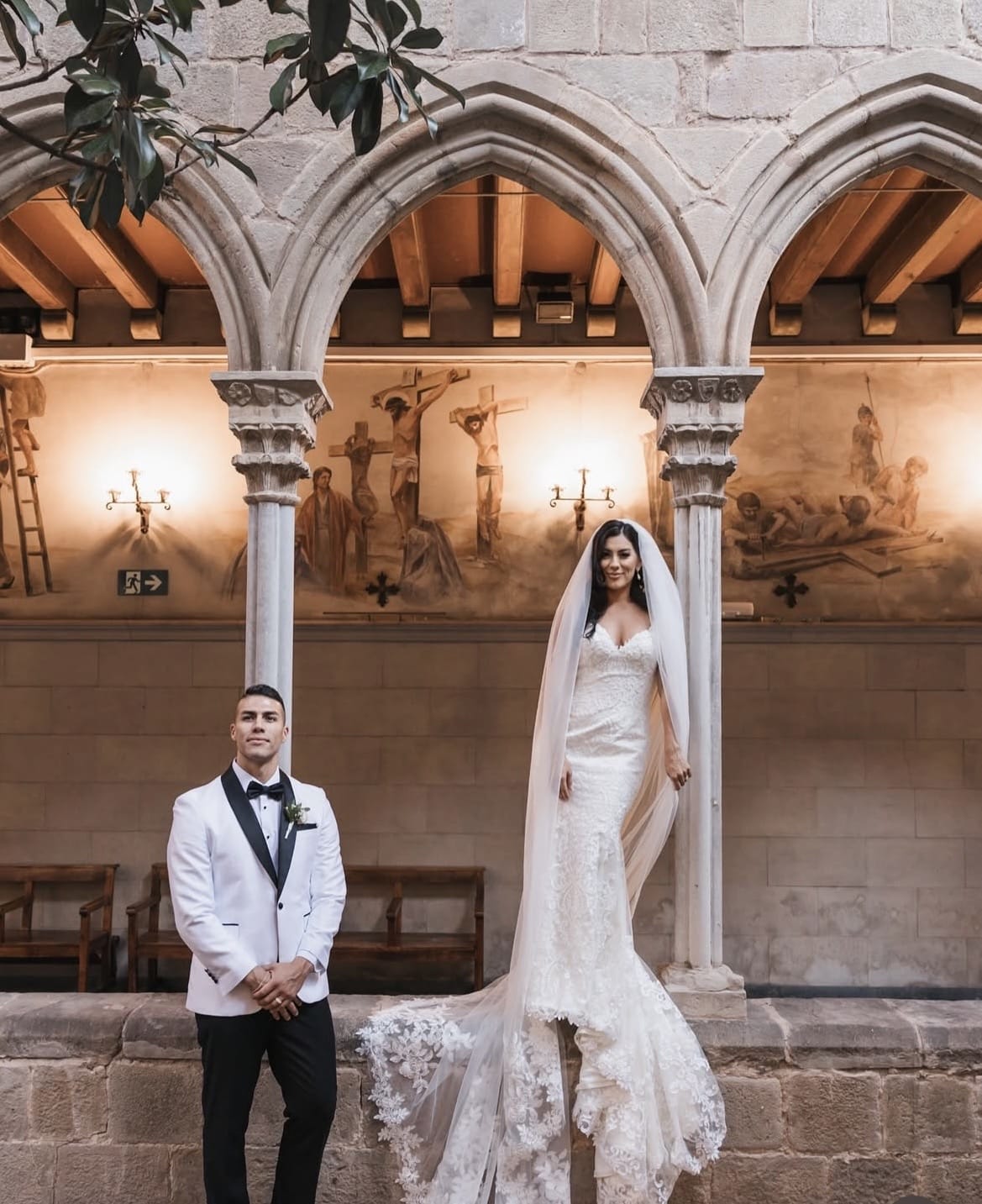 Novia con vestido de encaje y velo largo junto al novio en claustro histórico en Barcelona, maquillaje natural y luminoso